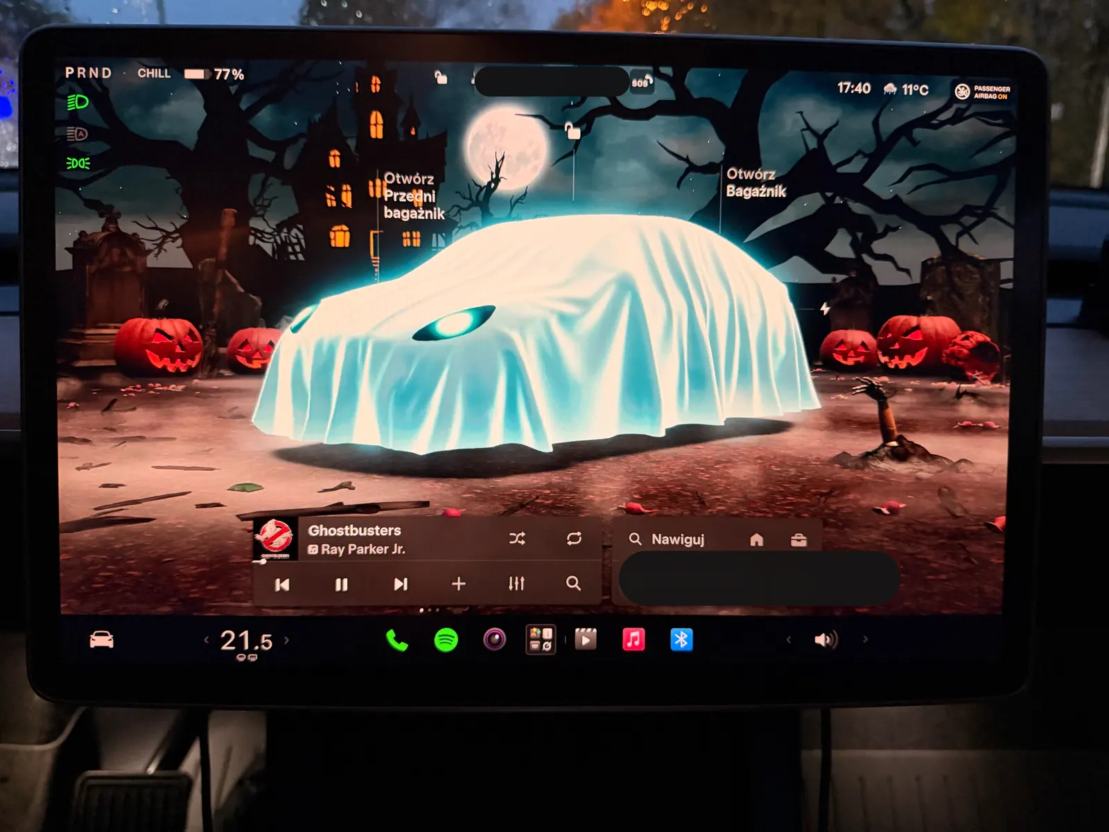 Tesla Halloween Mode parked scene: a sheet-covered ghost car under a full moon, with a haunted house and pumpkins